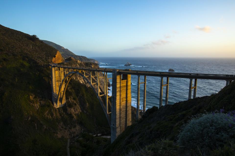 The setting sun falls on an old bridge near ocean cliffs.