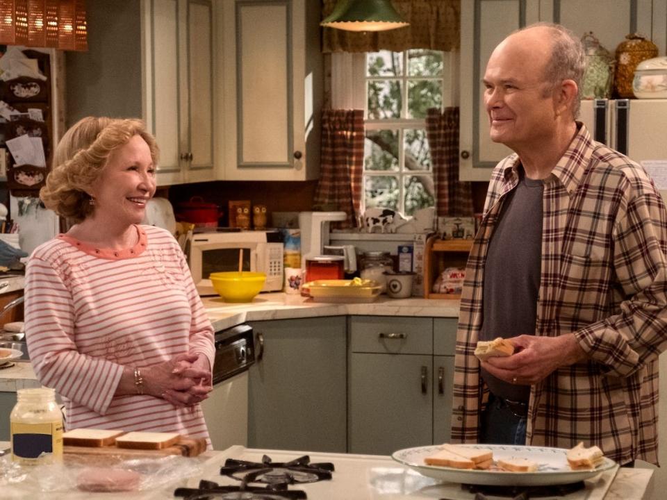A older woman and man stand smiling in a kitchen on the set of a sitcom TV show.