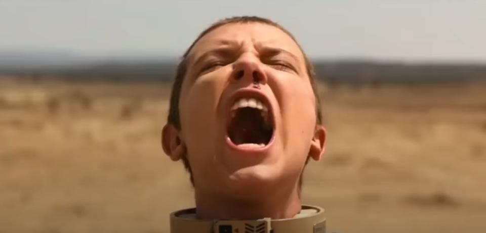 A girl with shorn hair wearing a bomb collar screams expressively with her eyes shut and her nose bleeding