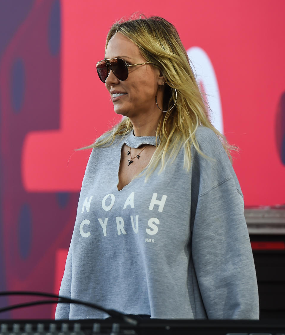 Woman in sweatshirt with "Noah Cyrus" text, standing with microphone, outdoors