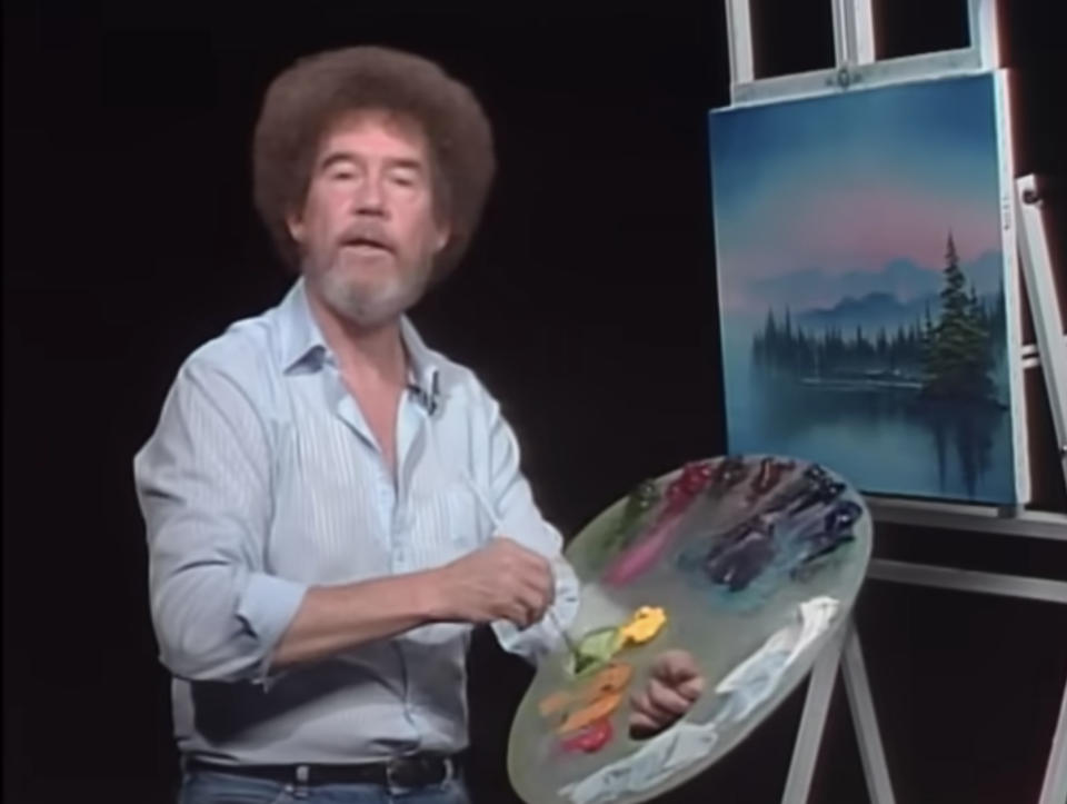 Bob Ross painting a serene lake scene on canvas, holding a palette with various paint colors while wearing a striped shirt