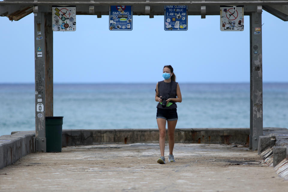 A woman wears a mask as she walks along a closed Waikiki Beach pier in Honolulu on Saturday, March 28, 2020. Like many cities across the world, Honolulu came to an eerie standstill this weekend as the coronavirus pandemic spread throughout the islands. But Hawaii officials went beyond the standard stay-at-home orders and effectively flipped the switch on the state's tourism-fueled economic engine in a bid to slow the spread of the virus. As of Thursday, anyone arriving in Hawaii must undergo a mandatory 14-day self-quarantine. The unprecedented move dramatically reduced the number of people on beaches, in city parks and on country roads where many people rely on tourism to pay for the high cost of living in Hawaii. (AP Photo/Caleb Jones)