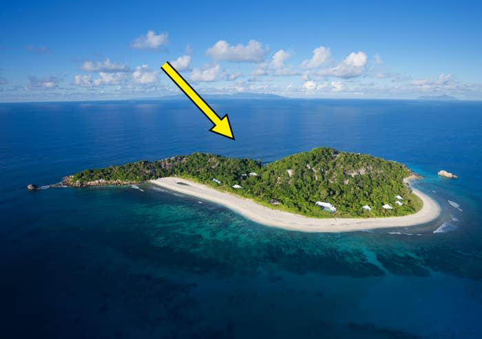 Aerial view of a small, lush island surrounded by sandy beaches and clear blue ocean waters. The island has several small buildings scattered along the shoreline