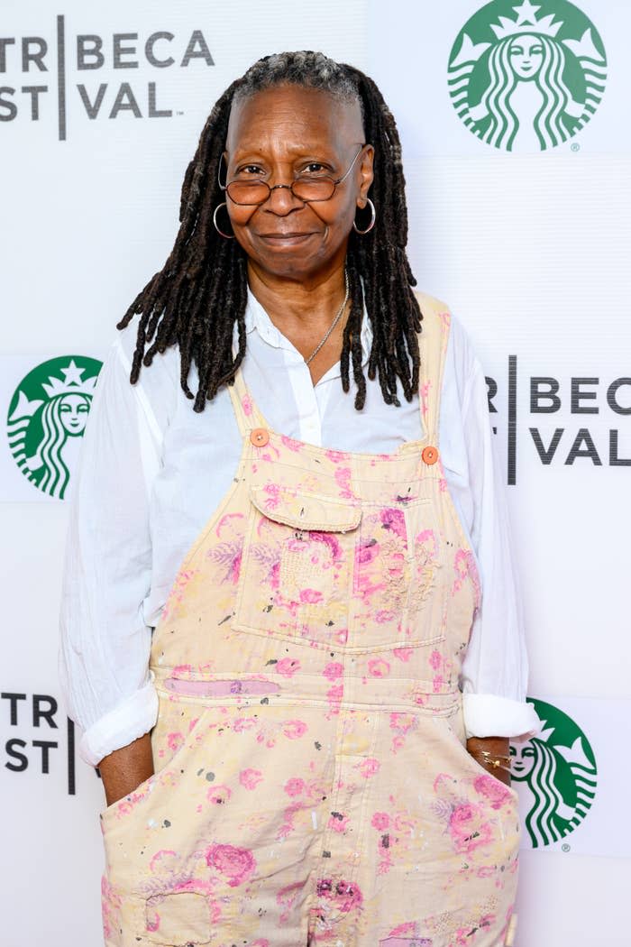 Whoopi Goldberg at an event, wearing pink paint-splattered overalls over a white shirt, with a Tribeca and Starbucks logo backdrop