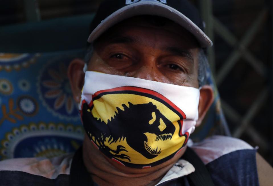 El guardia nocturno Félix Ortiz usa una máscara con el logotipo de Jurassic World mientras posa para una foto frente a una tienda cerrada por la cuarentena para evitar la propagación del nuevo coronavirus en Asunción, Paraguay, el jueves 30 de abril de 2020. Ortiz también vende máscaras como la que lleva puesta. (AP Foto / Jorge Sáenz)