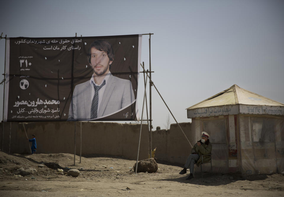 In this Wednesday, March 19, 2014 photo, a police mans a traffic checkpoint next to an election poster showing provincial candidate Mohammad Harun Mosawer on the outskirts of Kabul, Afghanistan. Wardak pulled out of the presidential elections but warlords, with a violent past have played a role in influencing Afghan politics since a U.S.-led coalition helped oust the Taliban in 2001. But they are emerging to play an overt political role in next month’s presidential elections as President Hamid Karzai leaves the scene. (AP Photo/Anja Niedringhaus)