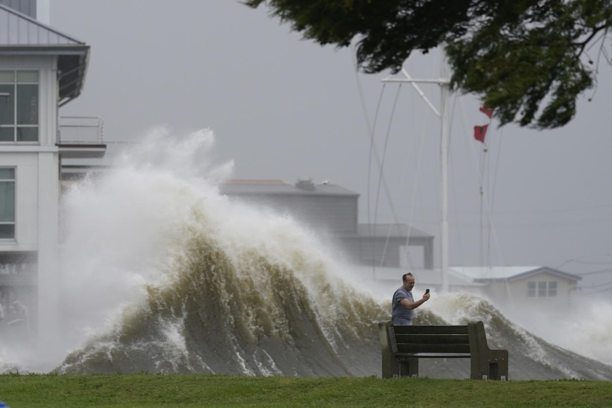 A man holds a phone near an urban coastline as a large wave swells behind him.
