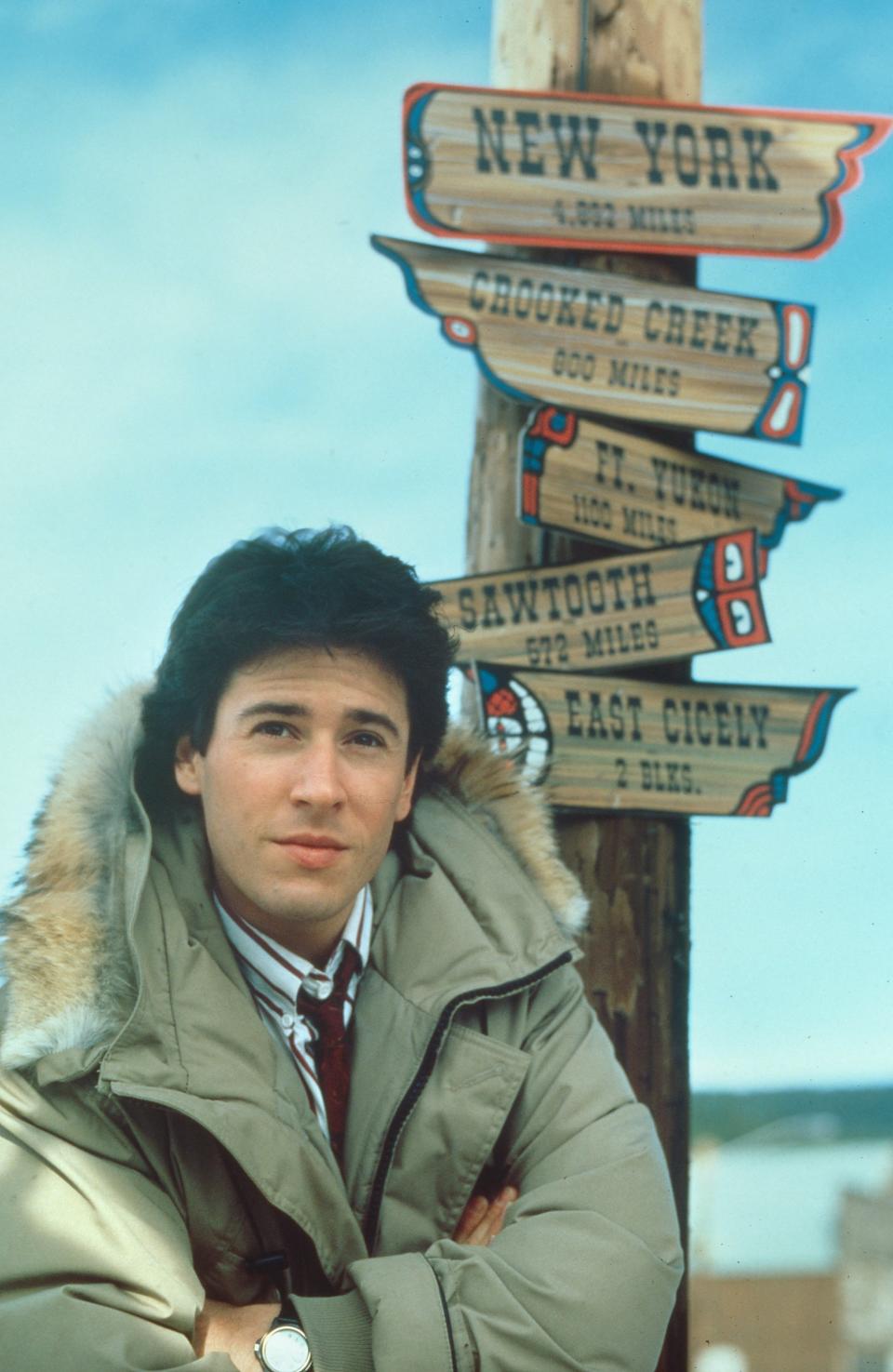 Man in parka by signpost with directional signs; relevant for "Northern Exposure" TV show