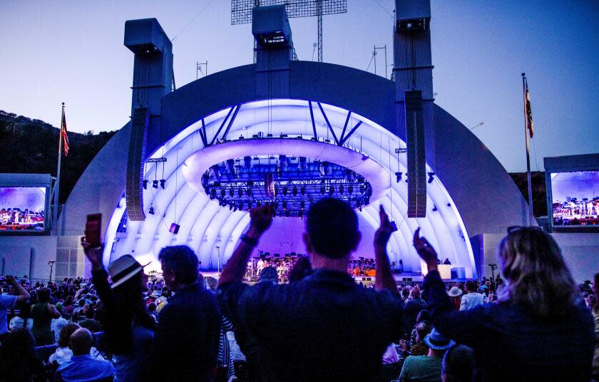 Silhouettes of concert attendees raising their hands in front of the Hollywood Bowl, which has a purple hue