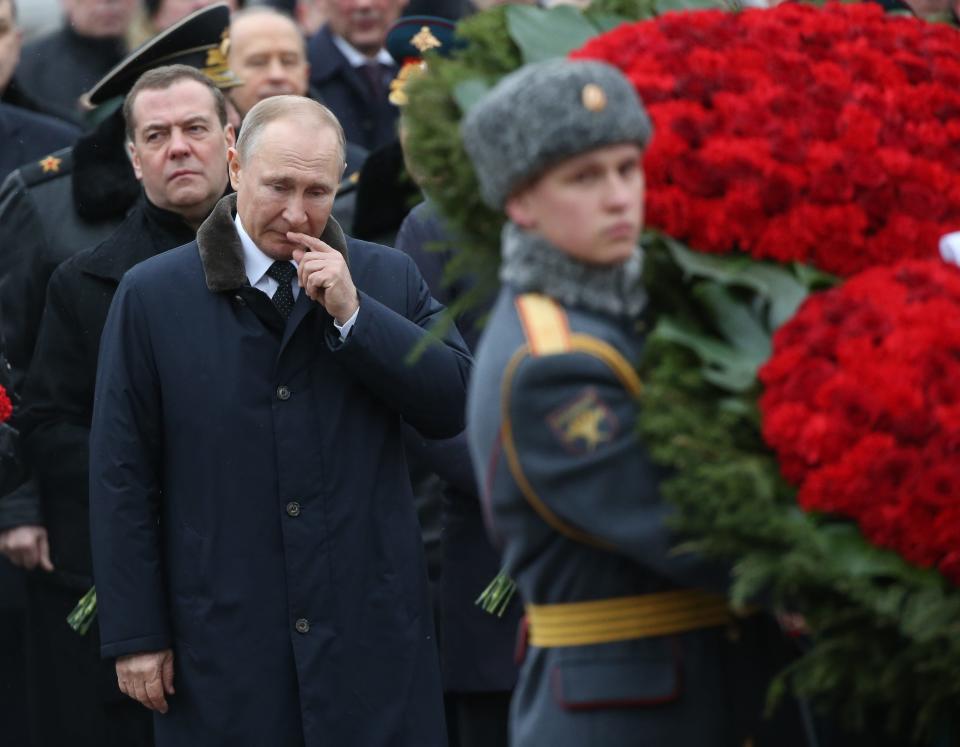 Dmitry Medvedev standing behind Vladimir Putin, with a soldier holding a wreath of red flowers in the foreground.