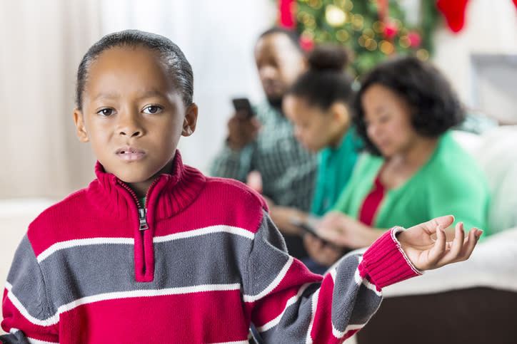 Child in a sweater stands perplexed while adults in the background use technology, possibly exploring work-from-home dynamics