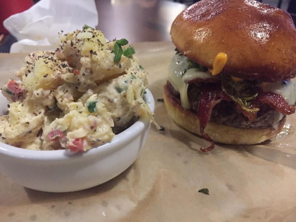 The Abridged Burger is a 7-ounce, all-beef ground-brisket patty topped with white American cheese, applewood-smoked bacon, caramelized red onions, fried Brussels sprouts petals and house aioli. Served on a brioche bun. Potato salad as a side item.