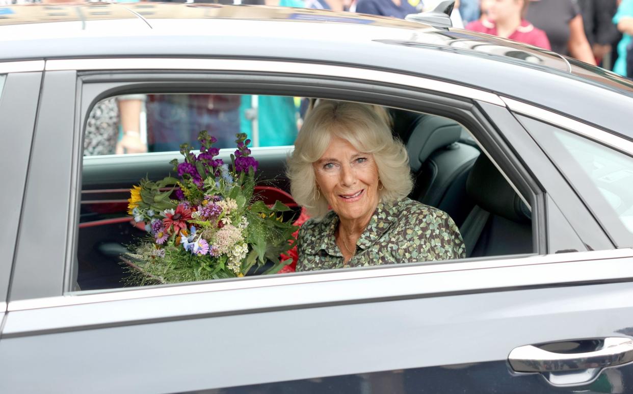 Queen Camilla sits in the back of a car with a bouquet of flowers, smiling