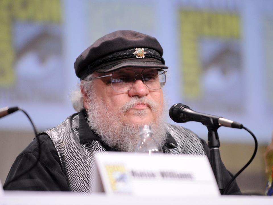 george r.r. martin sitting on a comic con panel, wearing a black hat and with a microphone in front of him