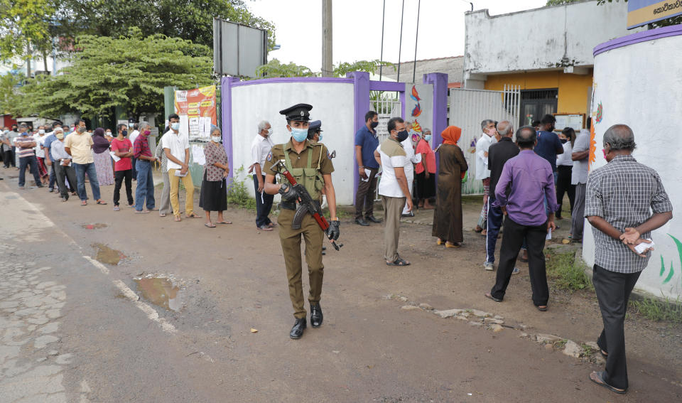 Sri Lankans stand in a queue as a police officer stands guard outside a polling station in Colombo, Sri Lanka, Wednesday, Aug. 5, 2020. Sri Lankans started voting Wednesday to elect a new Parliament that is expected to give strong support to the powerful and popular Rajapaksa brothers. (AP Photo/Eranga Jayawardena)