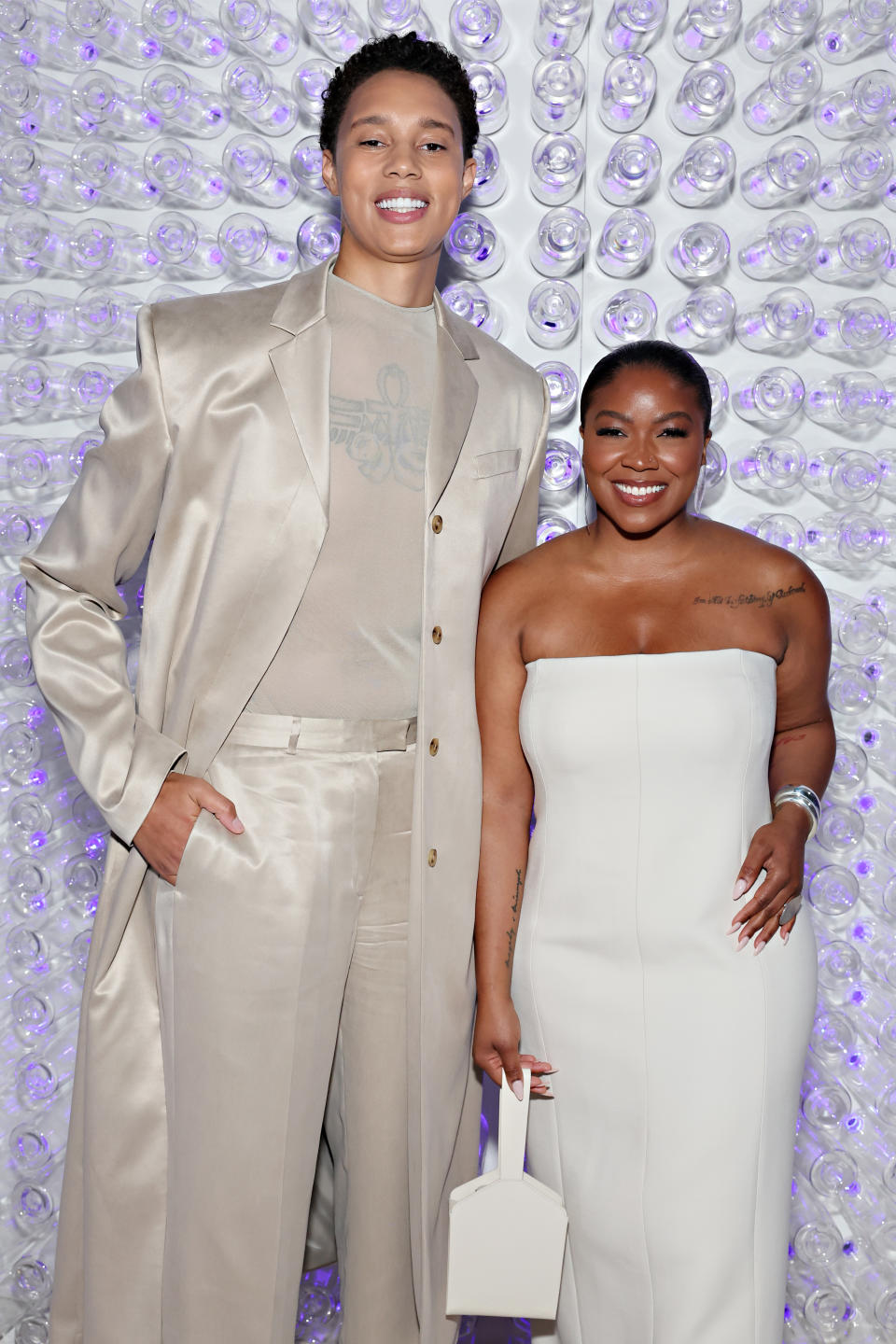 Brittney Griner in a silky suit and Cherelle Griner in a strapless dress smile for a photo against a backdrop of clear circular objects