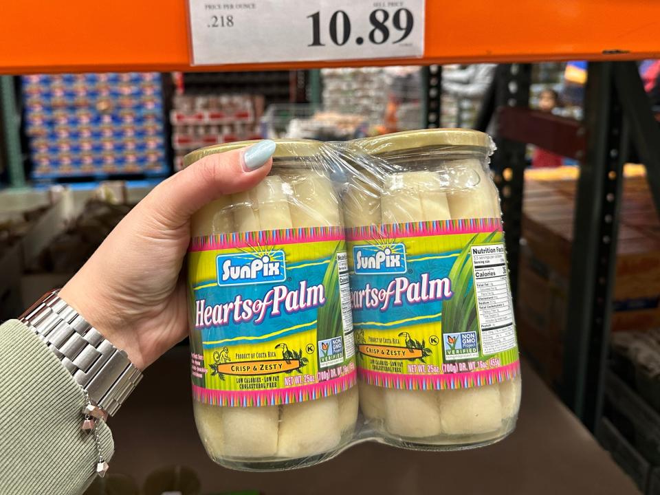 A hand holds a two-pack of hearts of palm in clear glass jars. The jars have yellow, blue, and pink labels on them