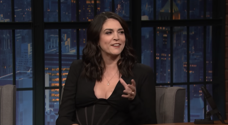 Cecily in a black outfit sitting, gesturing on the talk show