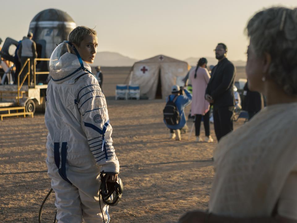 a woman in a spacesuit looks back at a crowd of others forlornly, walking across a brown field towards a red cross tent