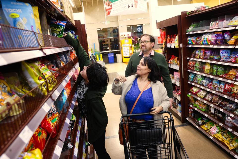 A teen reaches for snacks in the grocery store as his mother pushes a shopping cart and his father looks on.