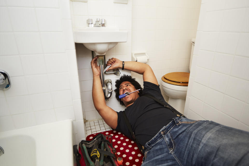 A guy on the floor underneath a bathroom sink working on it with a tool, holding a screwdriver in his mouth