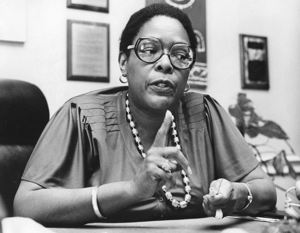 cardiss collins sits at a table and speaks while motioning with her hands, she wears a short sleeve shirt, glasses and jewelry