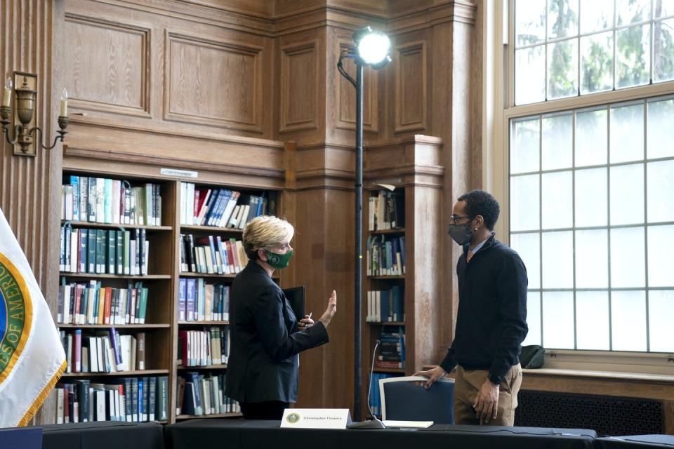 A masked woman and man speak under a spotlight in a booklined room