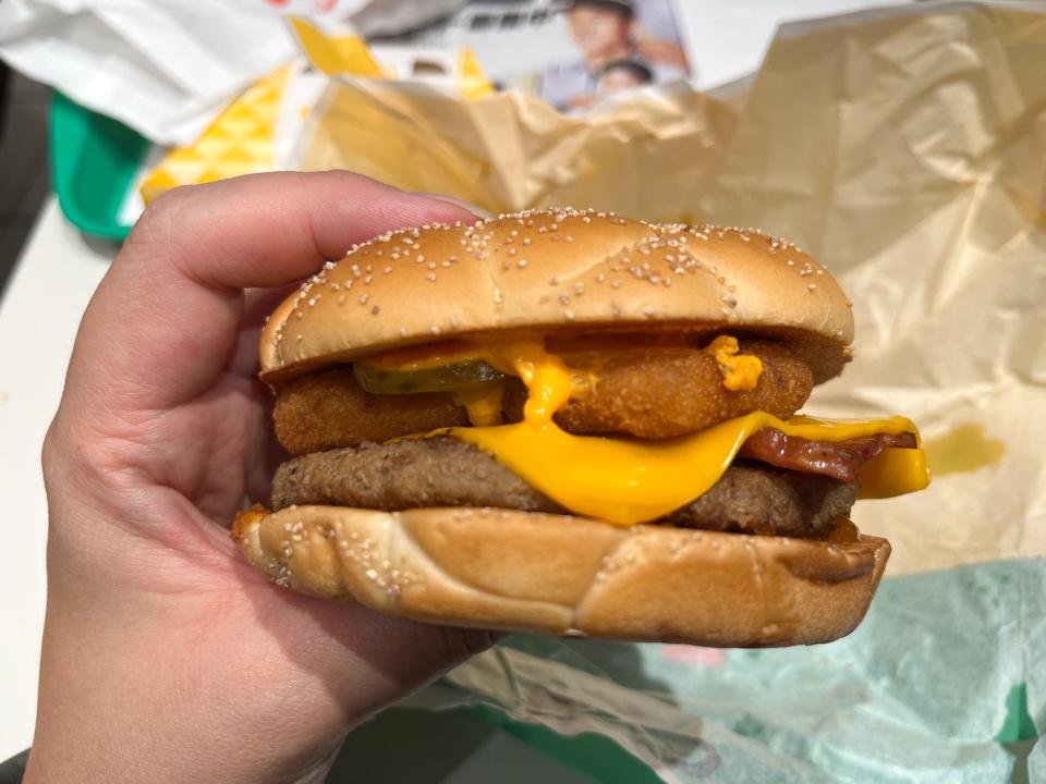 A close-up shot of a hand holding a fast food burger with onion rings and cheese.