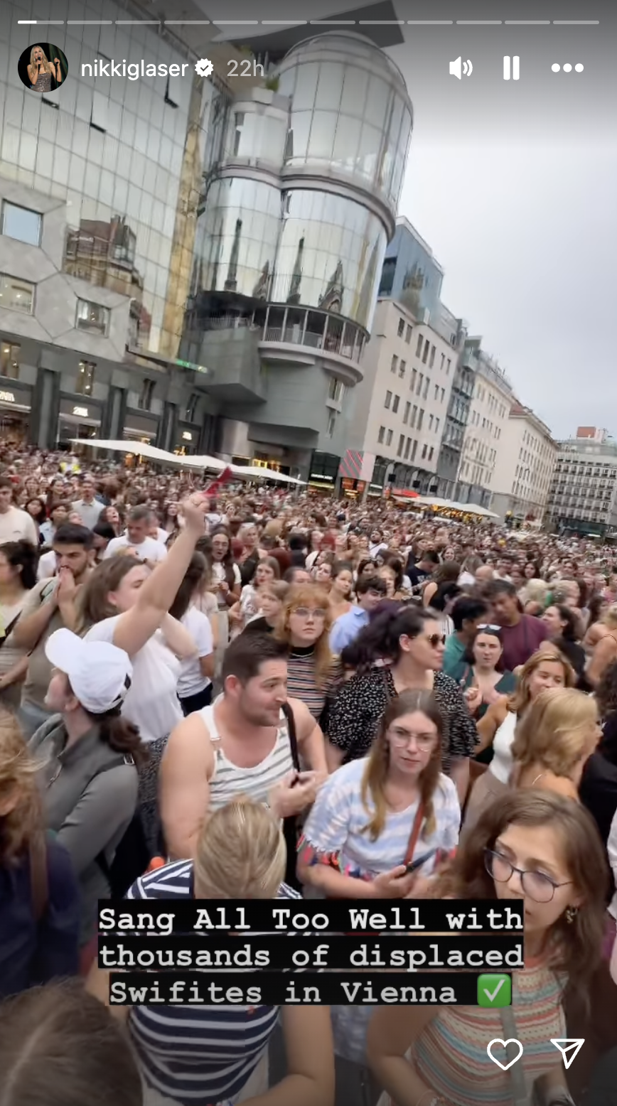 Hundreds of people crowd a city street with text superimposed that reads: Sang All Too Well with thousands of displaced Swifites in Vienna.