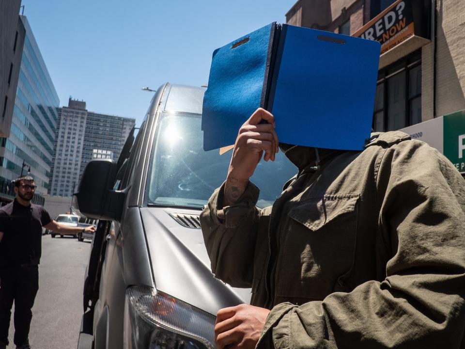 Flo Rida attempting to hide his face with an open blue binder as he walks in front of a Mercedes van.