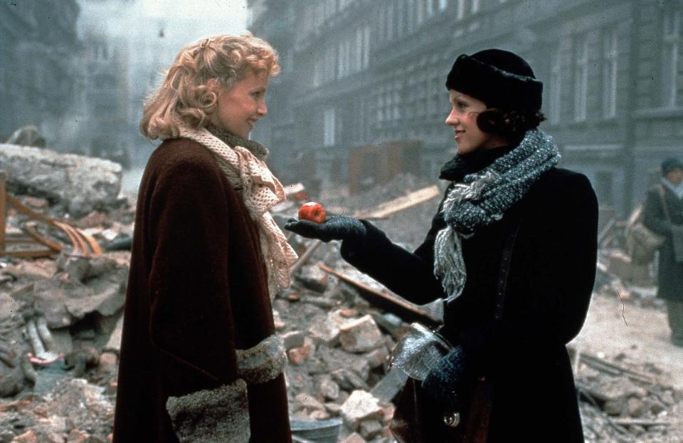 Anna (Susan Fleetwood) and Lili (Lisa Kreuzer) in winter coats and scarves, standing in a rubble-filled street scene from a WWII-era movie. Anna offers Lili an apple