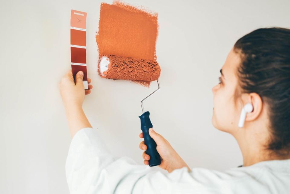 A woman holding a paint sample of orange shades against a white wall while painting with a roller.