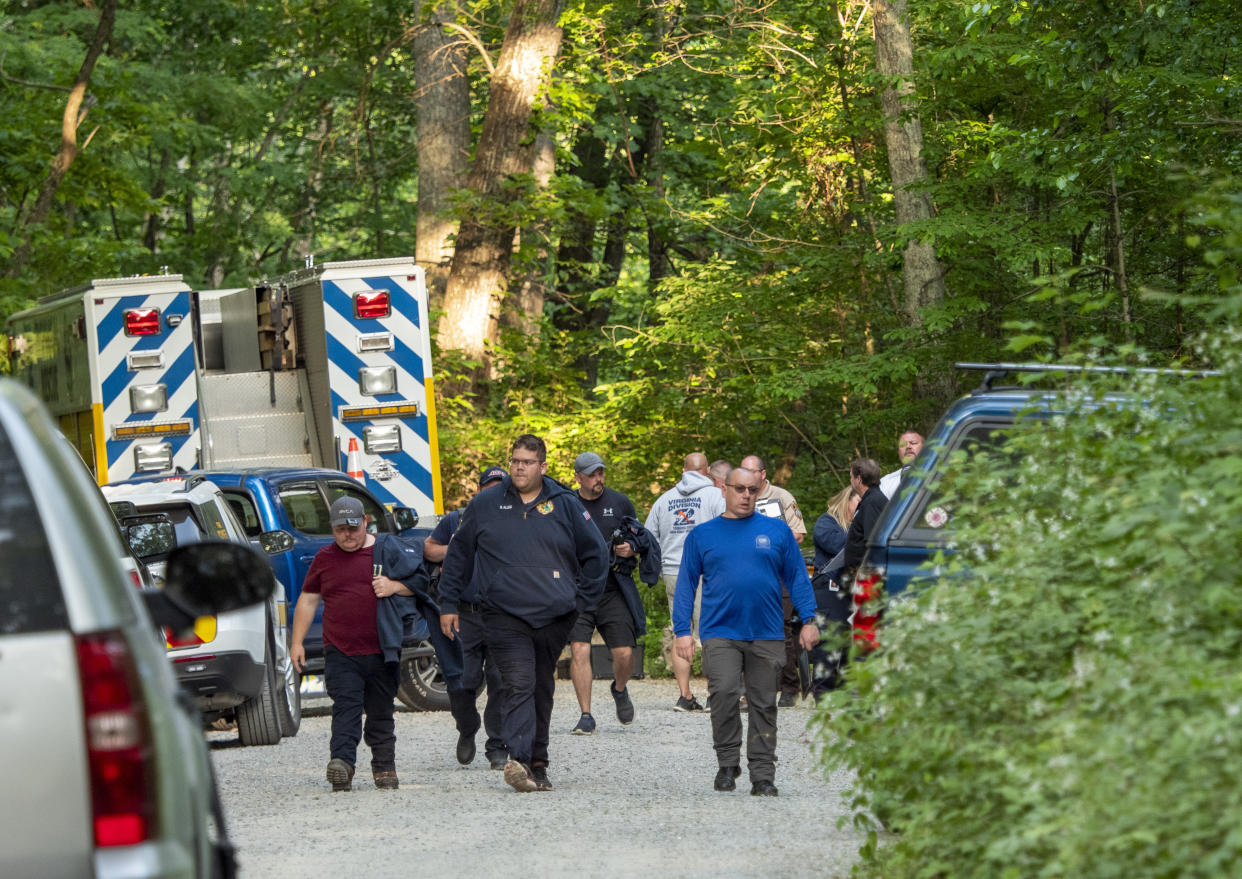 A dozen people walk or stand near parked cars and an emergency vehicle in what appears to be a clearing in the woods.