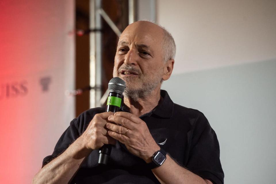 An older man in a black polo short speaks into a microphone.