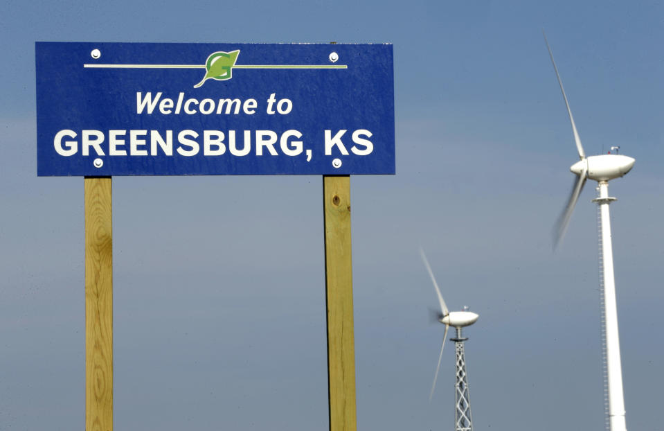 In this photo taken April, 18, 2014, wind turbines rise beyond a sign welcoming visitors to Greensburg, Kan. Seven years after an EF-5 tornado destroyed most of the community, Greensburg is one of the greenest towns in the country but struggles to regain residents that were displaced by the disaster. (AP Photo/Charlie Riedel)