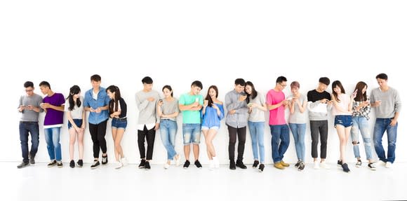 Wide angle shot of 17 male and female young Asians standing against a white background and looking at mobile devices.