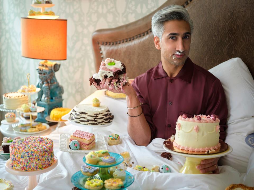 Tan France playfully eats cake, surrounded by cakes, in a white bed with brown backboard.