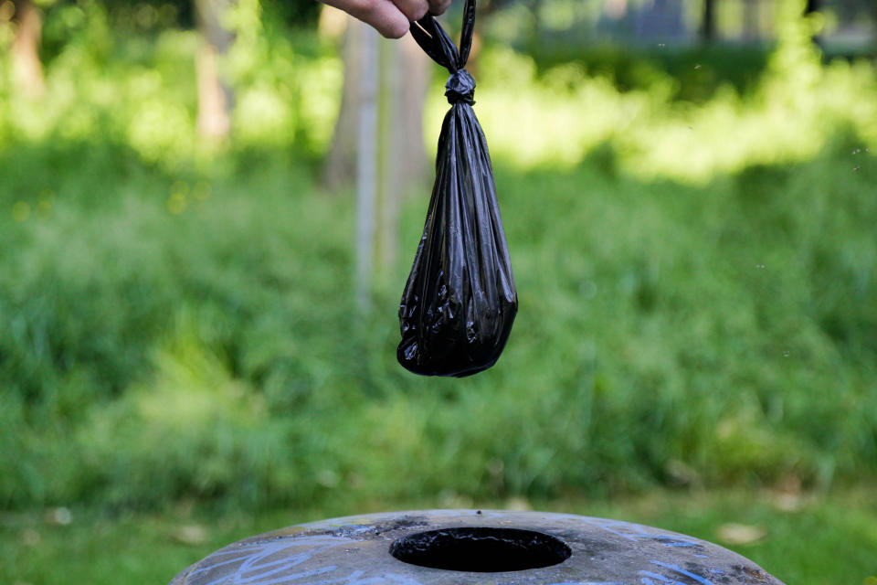 A hand holding a small black garbage bag over a trash can in an outdoor park setting