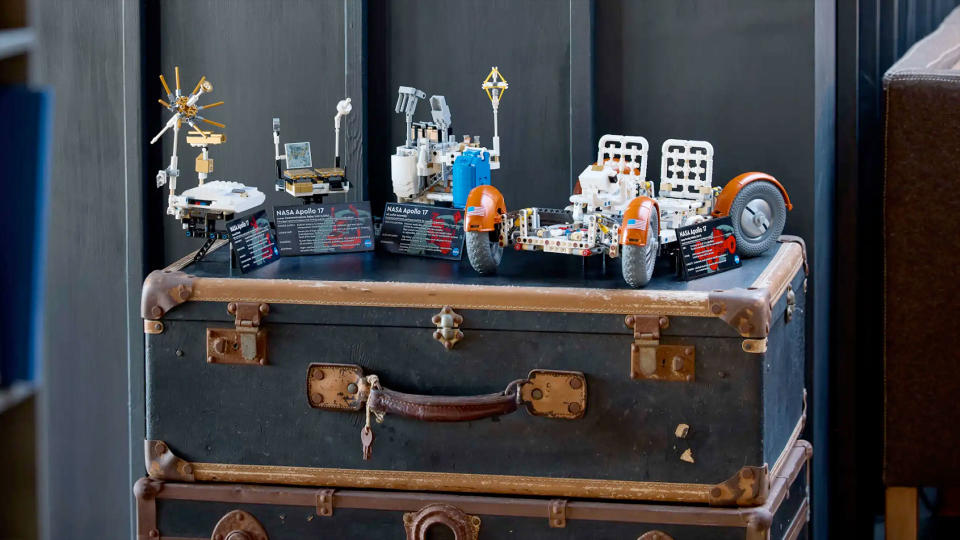 a white lego moon rover and several other small buildings sit atop a brown and black trunk against a gray wall.