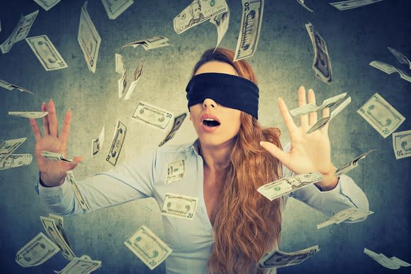 A blindfolded woman tries to catch paper money floating around her.