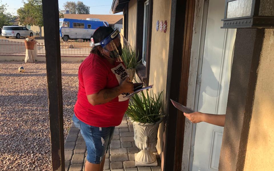 A well-built Latino woman with tattooed arms, wearing a red T-shirt that says "ONE JOB IS ENOUGH" and protective face masks, stands on a doorstep, leaning slightly forward as she speaks to an unseen voter. All that can be seen other person is a hand reaching out of the door, holding a pamphlet they have just been given. In the background is a gravel yard, and beyond that low tan-coloured houses, with an old silver and blue caravan parked in front of one of them - Laurence Dodds/Telegraph