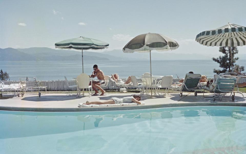 Umbrellas and sunbathers alongside a swimming pool. In the background, a span of blue water is backed by low mountains.