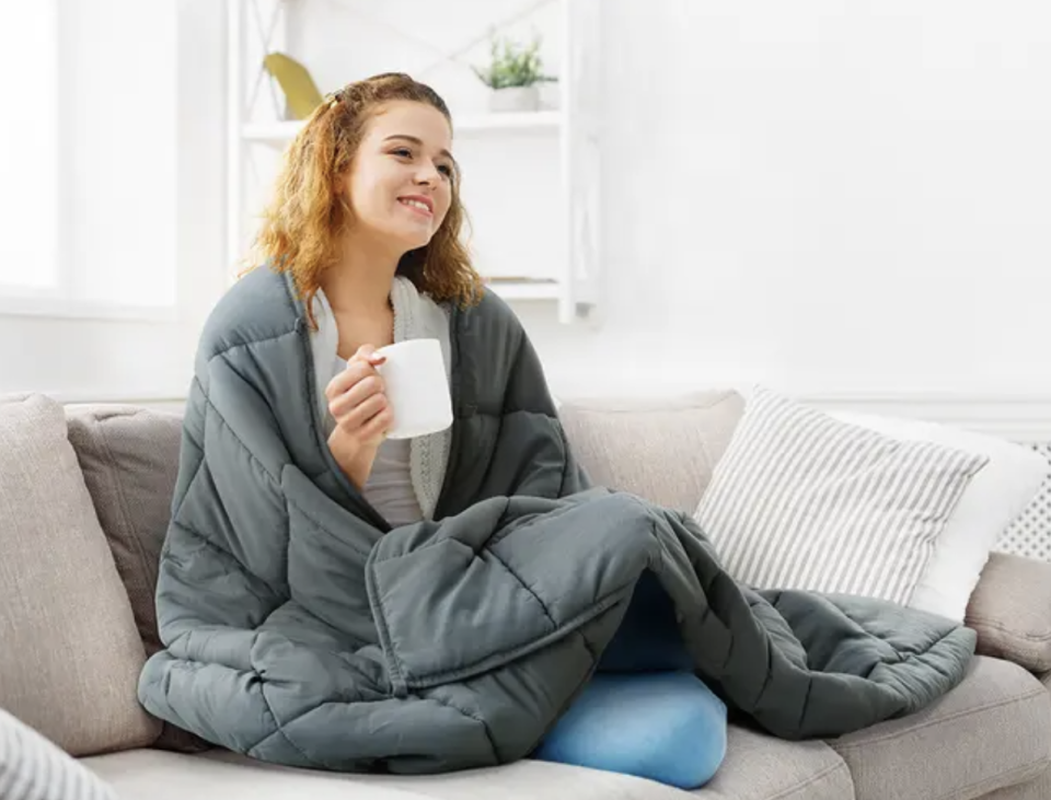 A woman sits on a grey lounge with striped cushions, holding a cup and smiling, wrapped in a grey Trafalgar All Seasons Weighted Blanket 9kg. Now $44.99