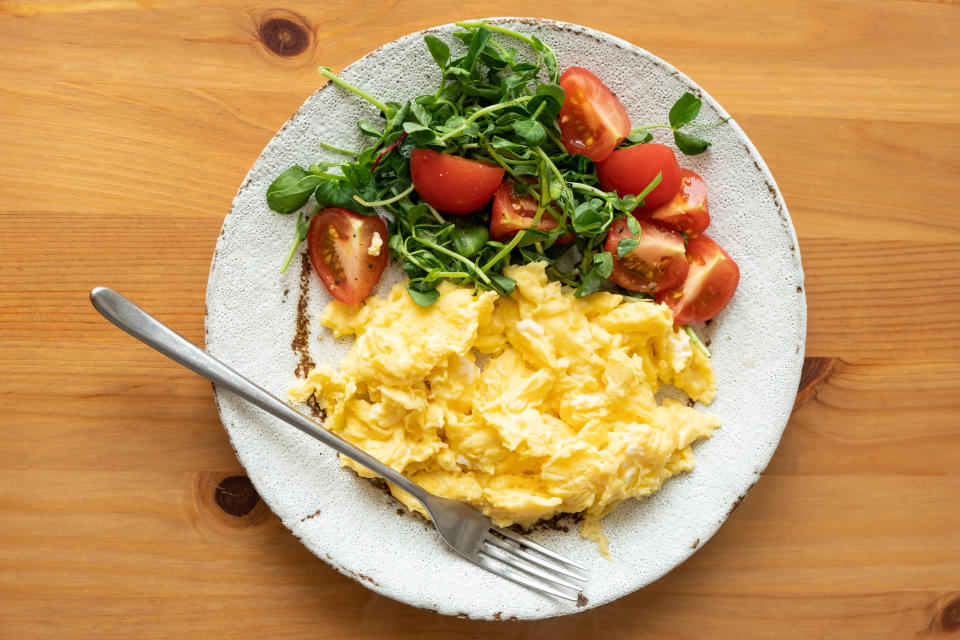 A plate with scrambled eggs and a tomato and arugula salad with a fork on the side