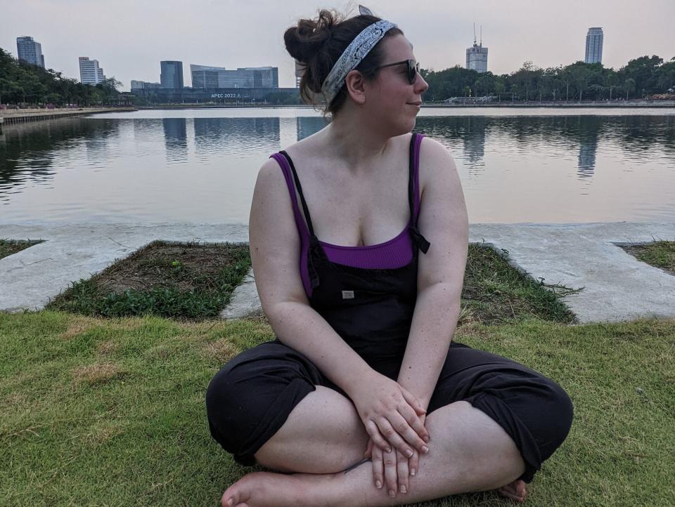 A woman sitting in a park cross legged in front of water and a skyline looking to the side.