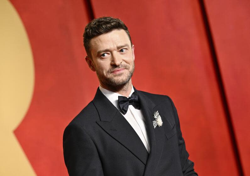 Justin Timberlake wears a black tuxedo while posing in front of a yellow and red backdrop