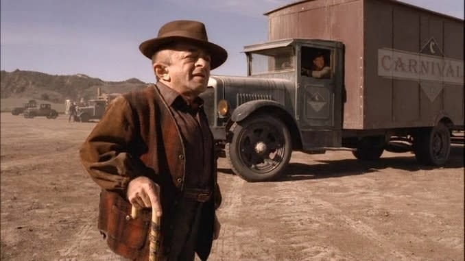 an older character with a cane and fedora outside of an old large carnival truck