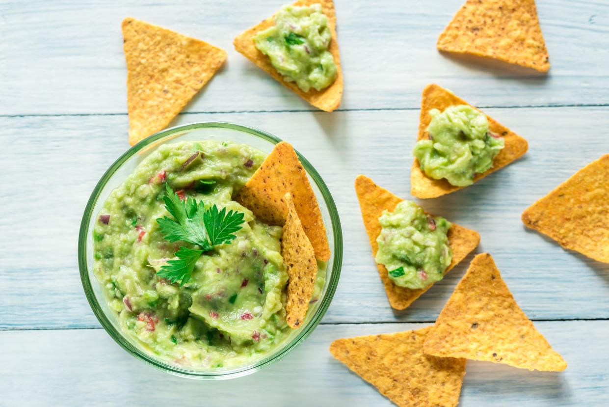 Easy guacamole in a glass bowl surrounded by tortilla chips, three with guacamole on a light blue wooden table