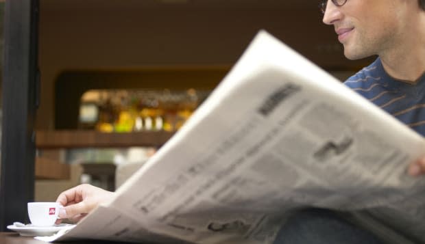 "Man sitting at cafe table reading newspaper, reaching for cup"