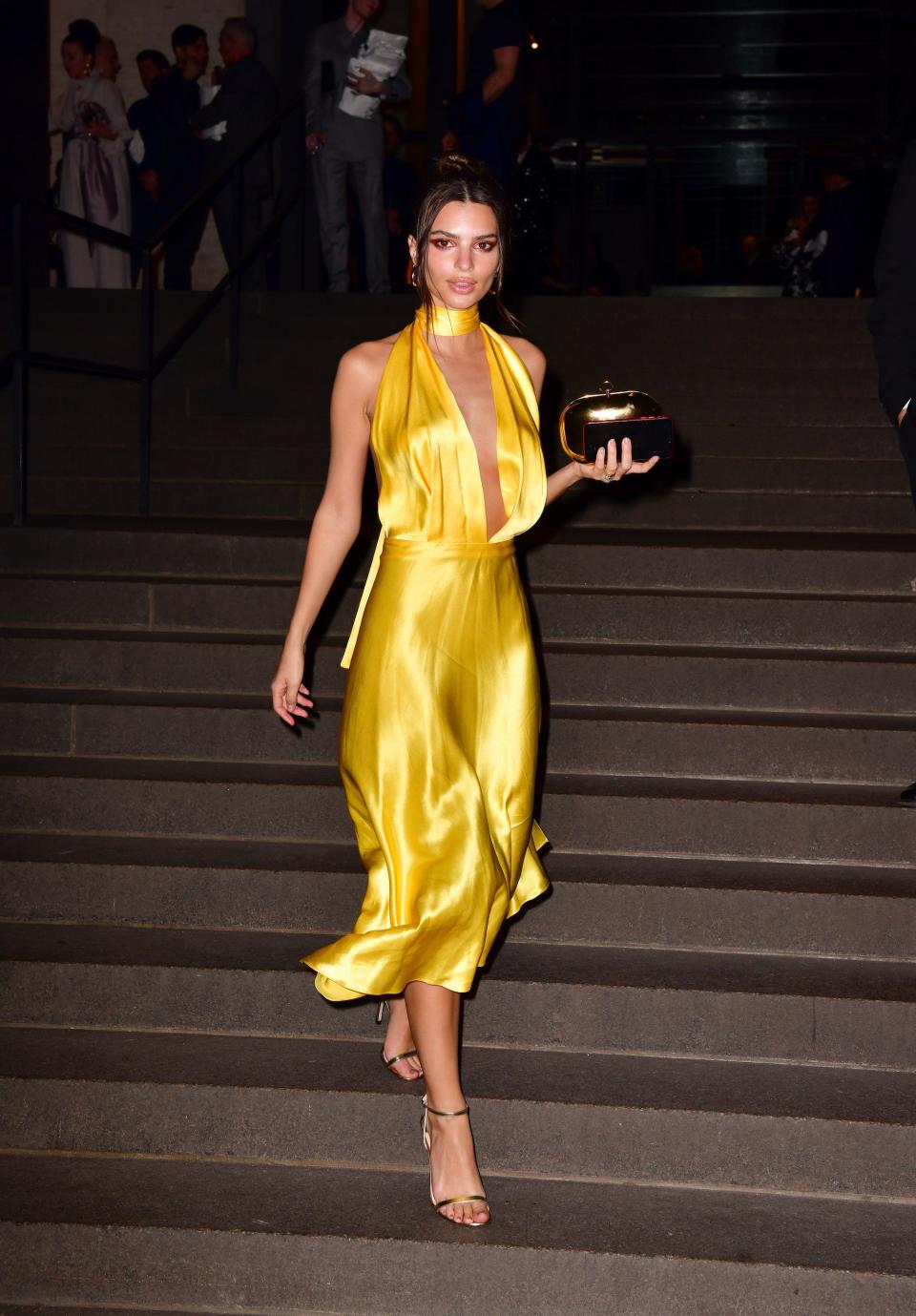 Emily Ratajkowski walks down a flight of stairs in a yellow dress.